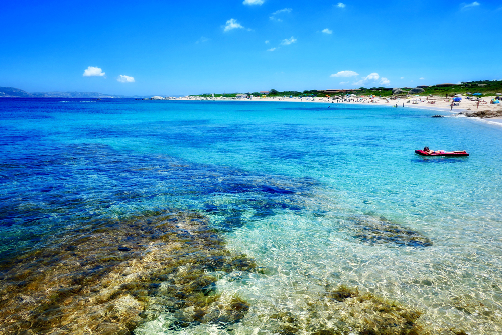 Eau turquoise en Sardaigne, femme sur matelas gonflable dans l'eau sur la droite et plage en arrière-plan. Gens sur la plage
